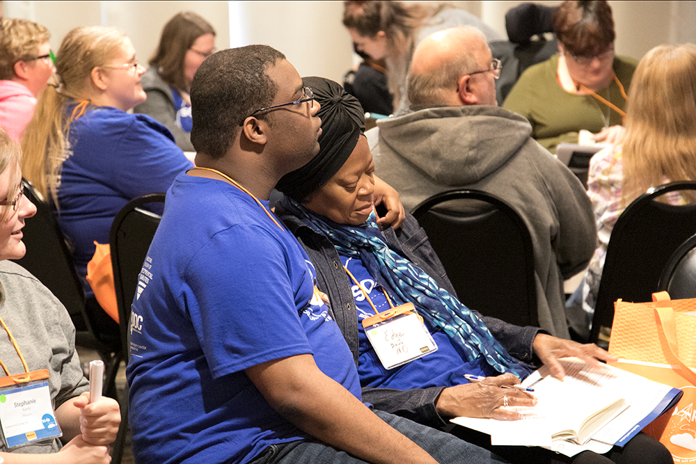 Mother and son conference attendees are photographed embracing one another as they listen to their breakout presentation speaker.