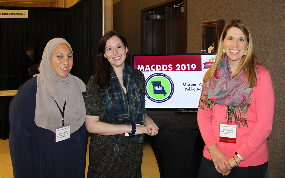 This photo shows IHD team members Cassie Webb, Megan LaMarche, and Jenny Turner standing and posing in front of a television screen that shows a MACDDS 2019 Banner.